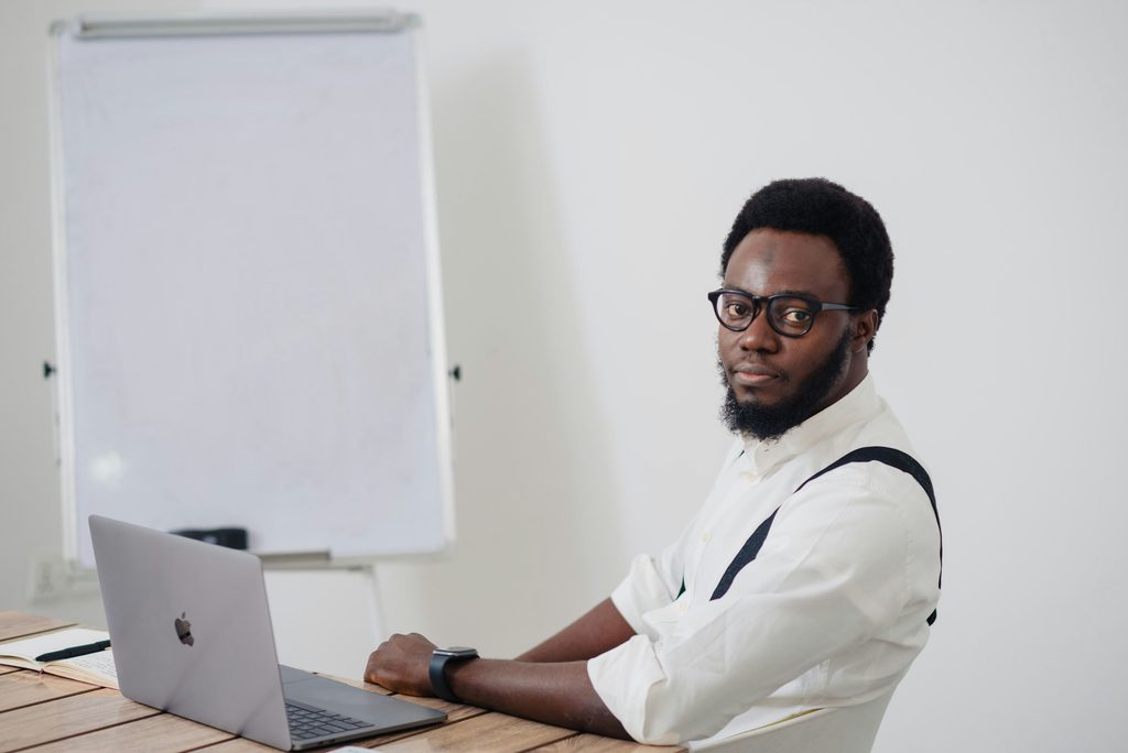 A man sits at a desk with an open laptop in front of him and a blank dry erase board off to the side.